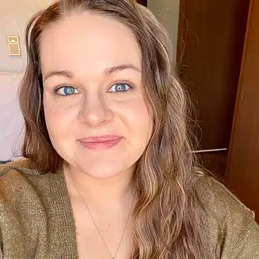 Selfie shot of a woman with curly hair, flashing a close-lipped smile while looking straight at the camera. Shot is slightly tilted to the left.