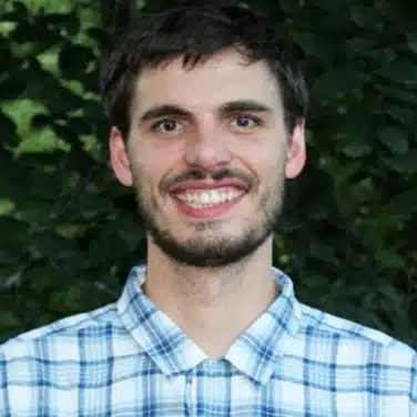 Headshot of a man wearing a collared shirt, flashing a smile while looking straight. Background is greenery.