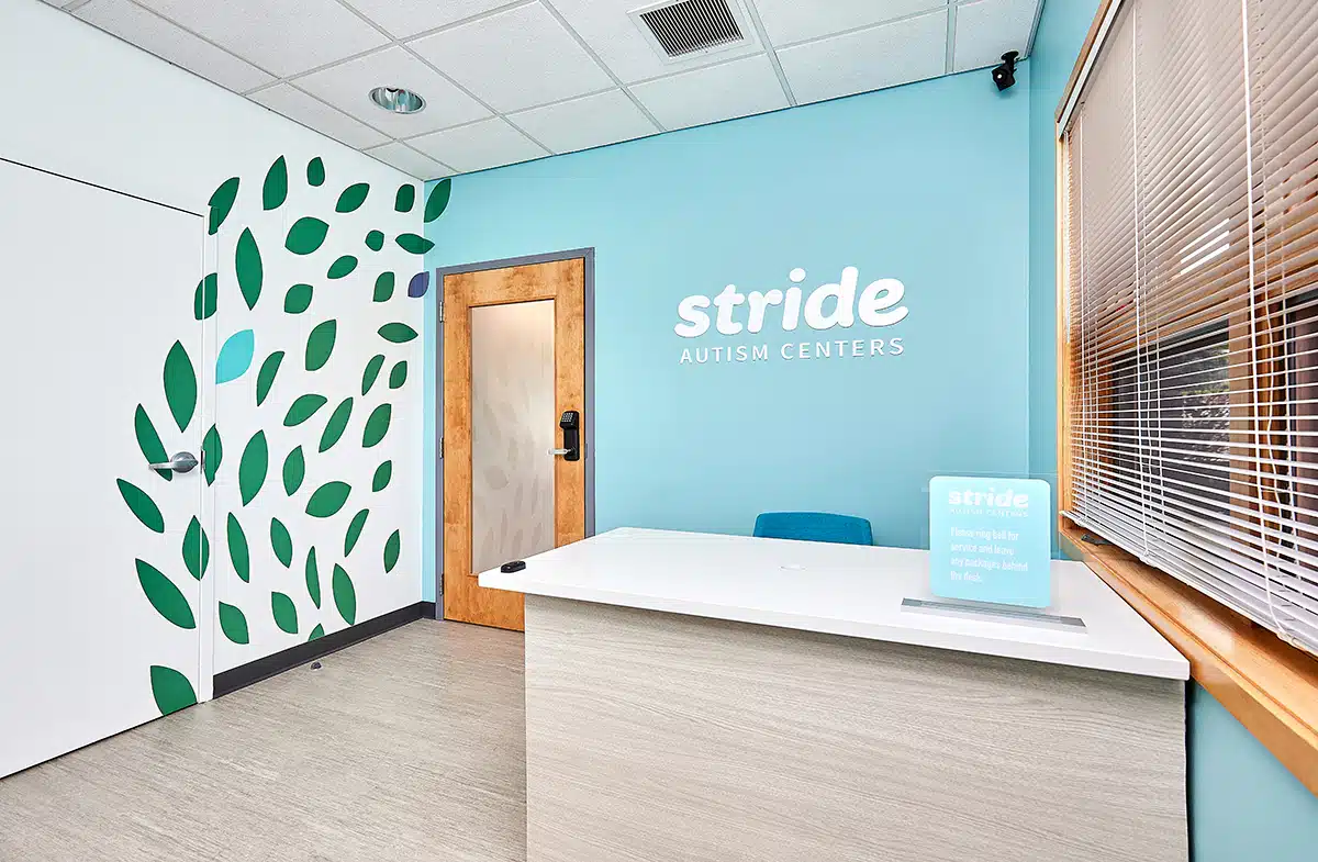 Photo of a reception area with a table and chair. The wall behind the chair has a signage that reads "Stride Autism Centers." There's also a wooden door and window with blinds.