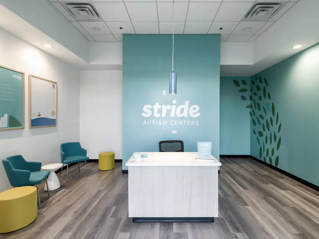 Photo of a room with a table and office chair at the center. Additional tables and chairs are on the left right. The text behind the central table and chair combo reads "Stride Autism Centers&reg;."