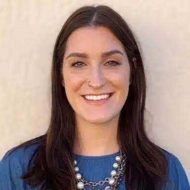 Headshot of a woman wearing necklaces, with slightly wavy hair, smiling while looking straight at the camera.