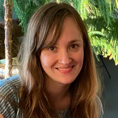 Selfie shot of a woman with wavy hair and bangs, flashing a smile while looking straight at the camera.