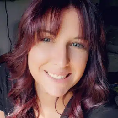 Selfie shot of a woman with curly hair and wispy bangs, smiling at the camera.