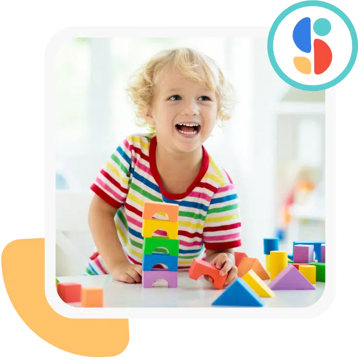 Photo of a child with curly hair. The child looks sideways while flashing a smile. The child is playing with colorful blocks and is wearing a striped shirt.
