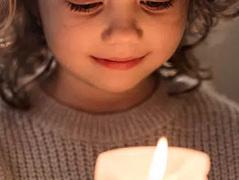 Photo of a child with curly hair looking at a lit candle while flashing a small smile.