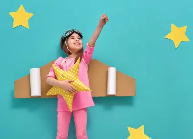 Photo of a girl with her left right outstretched upwards while her right hand holds a star-shaped pillow, Behind her is a cardboard shaped into airplane wings.
