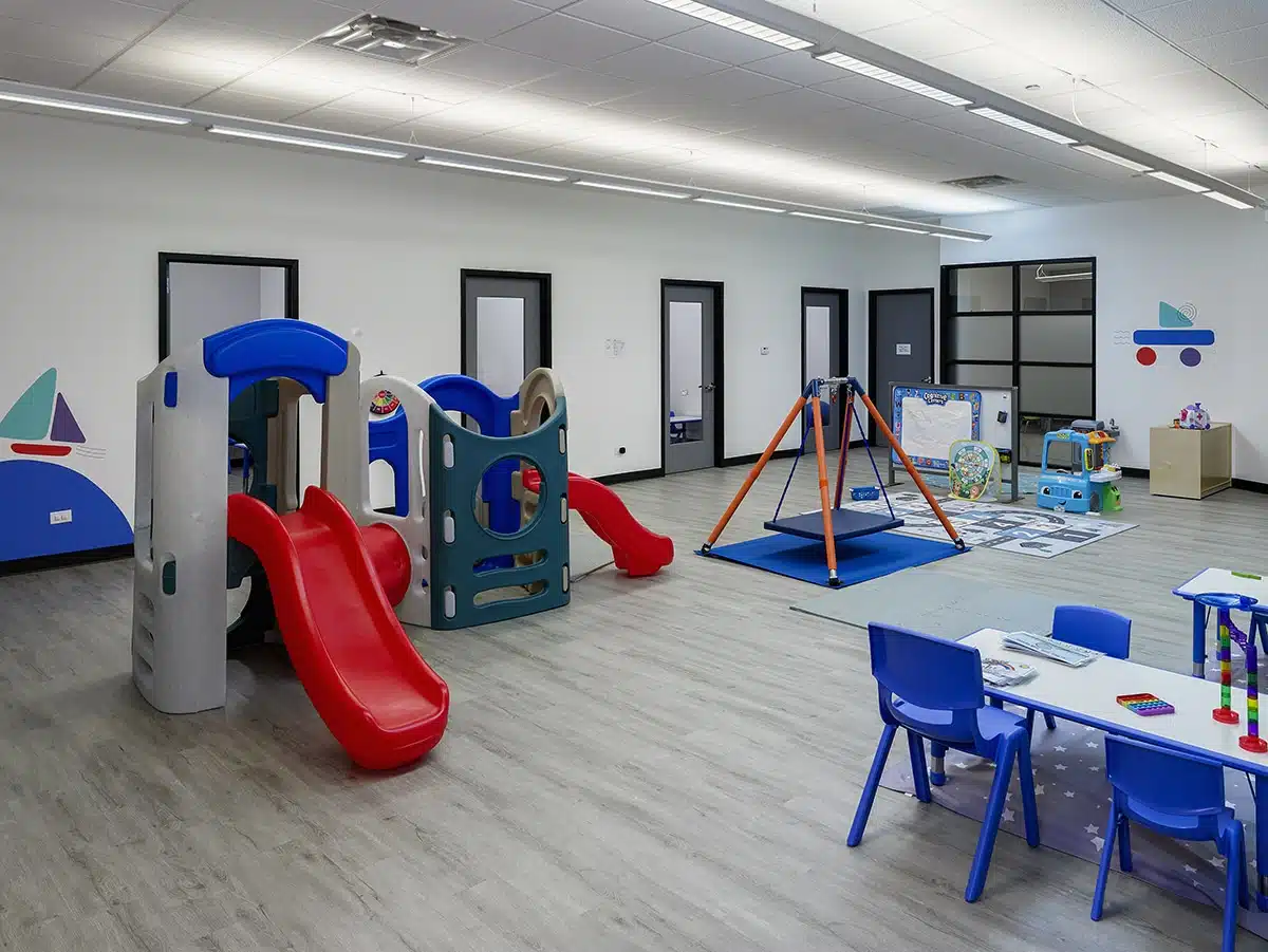 Photo of a play area with a swing, tables, chairs, shelves, slide, and a plastic car.
