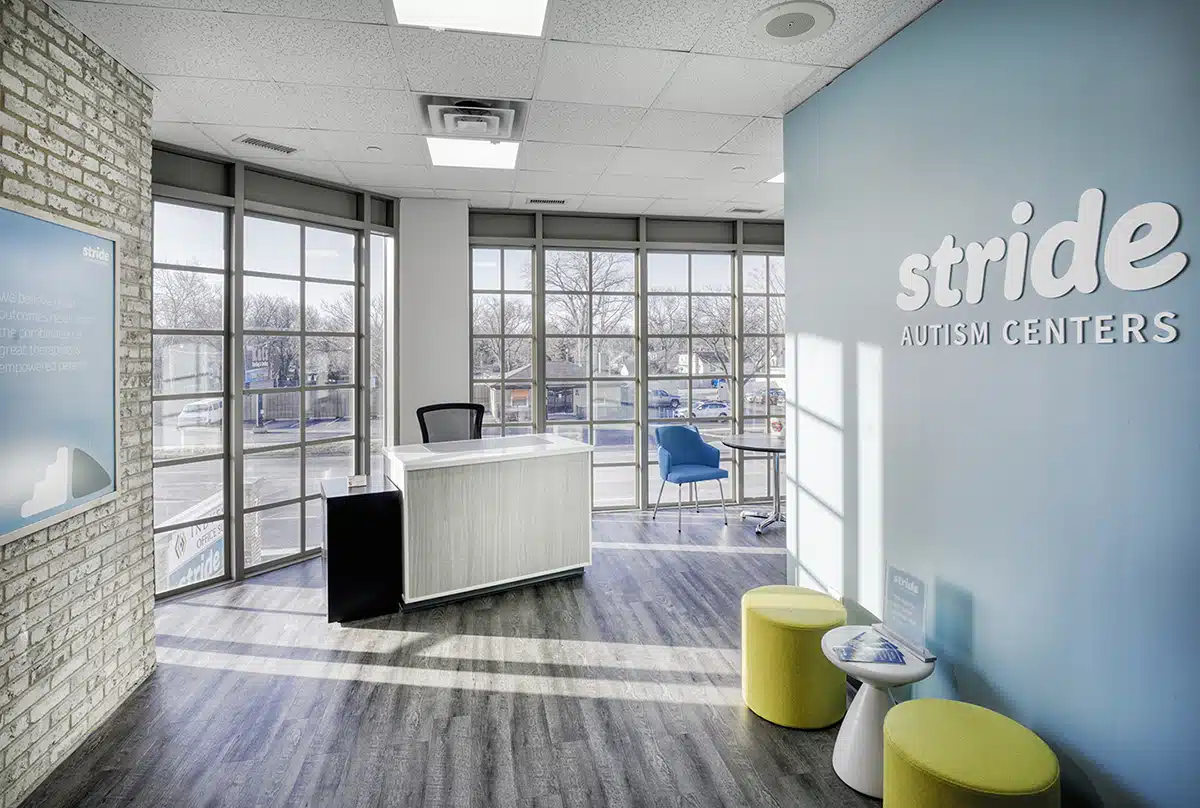 Photo of a room with glass panels, chairs, stools, tables. The wall on the right side has a signage that reads "Stride Autism Centers."
