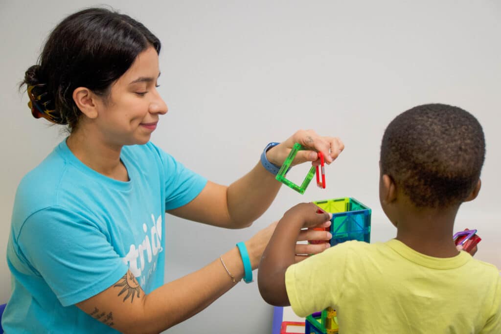 A clinician at Stride Autism Centers&reg; helps a young child build a tower using two colorful red and magnetic toy blocks.