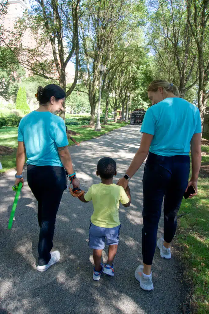 Dos terapeutas de Stride Autism Centers® pasean al aire libre, cogidos de la mano de un niño pequeño, por un sendero bordeado de árboles.