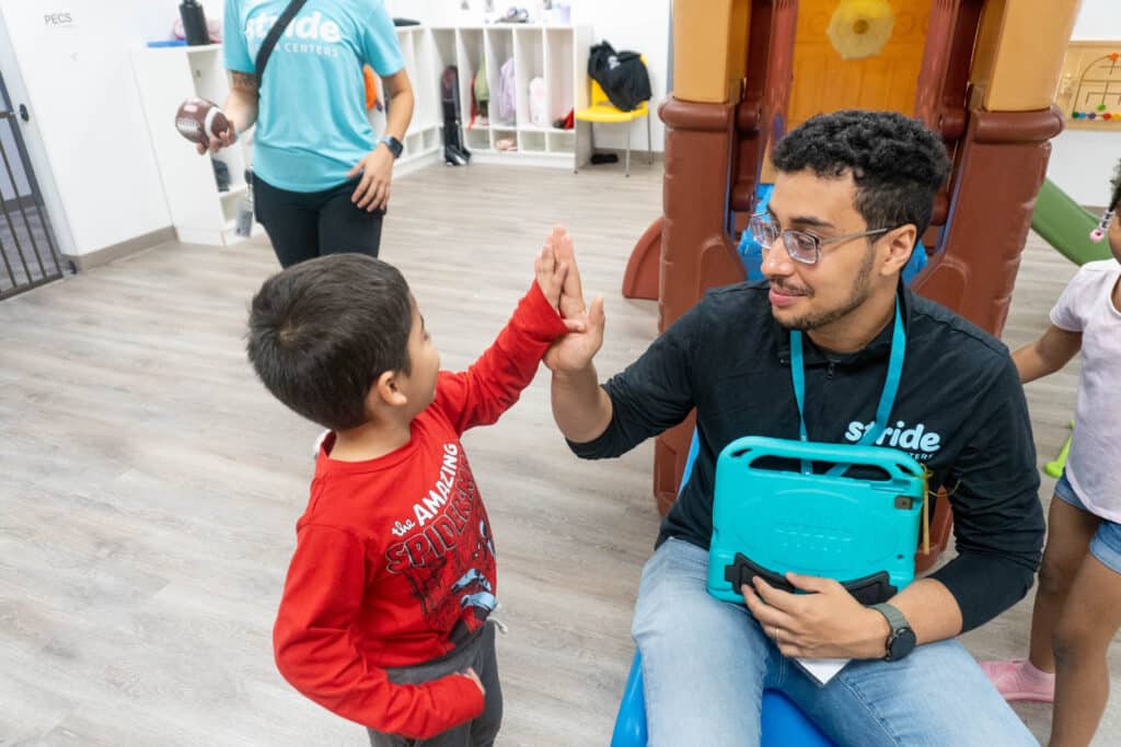 A Stride Autism Centers&reg; clinician wearing glasses giveing a high five to a child in a red hero shirt in a playroom.