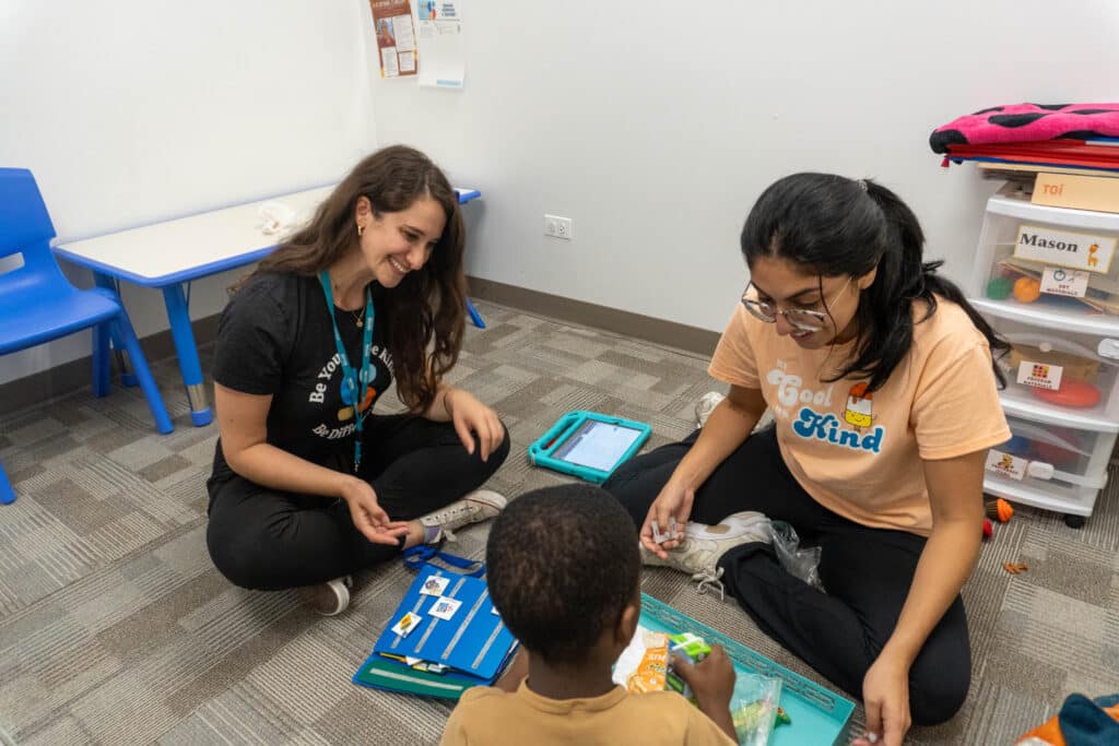 Two smiling Stride Autism Centers&reg; staff sit on the floor with a child using icons during a pediatric ABA therapy session