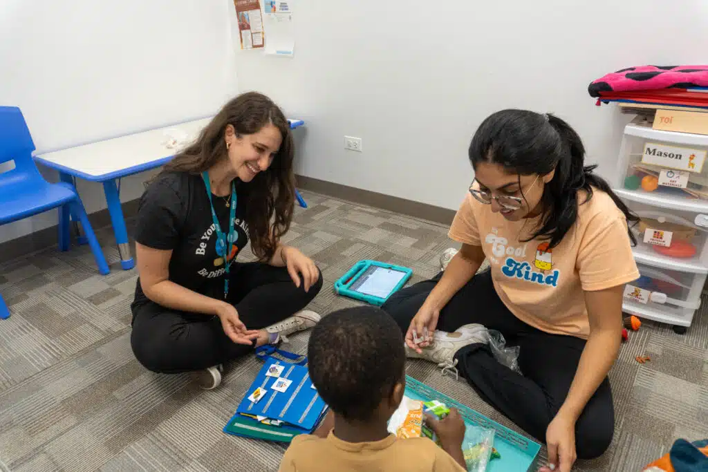 Two smiling Stride Autism Centers&reg; staff sit on the floor with a child using icons during a pediatric ABA therapy session